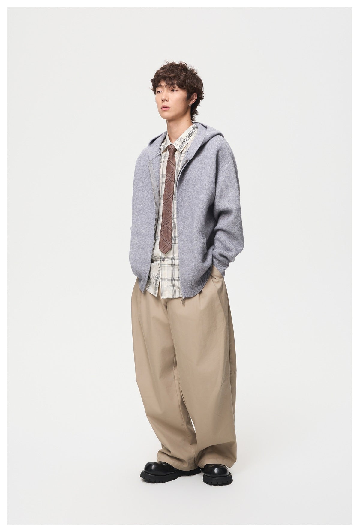 Full-length model wearing a light gray zip-front hooded knit jacket with a plaid shirt, tie, and beige trousers.
