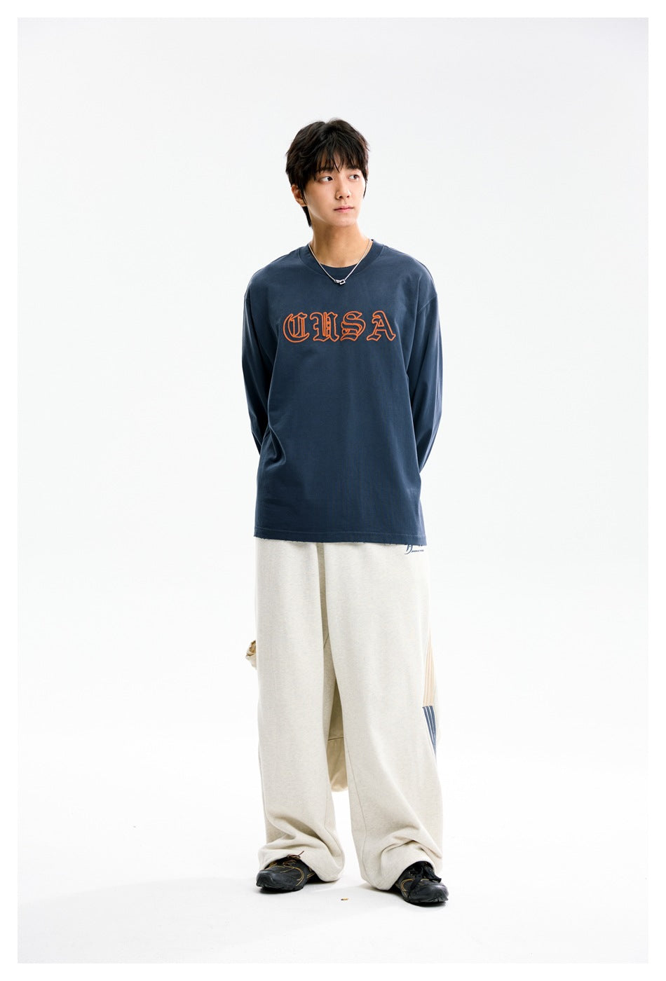 Model in a blue long-sleeve top with orange CUSA embroidery and light wide-leg pants, looking to the side with hands behind