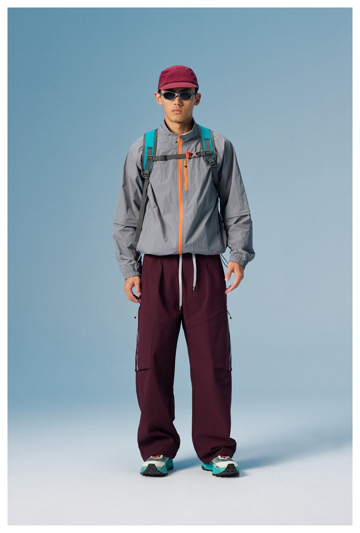 Full-length front view of a model in a gray jacket with orange zips, burgundy cargo pants, and a backpack.