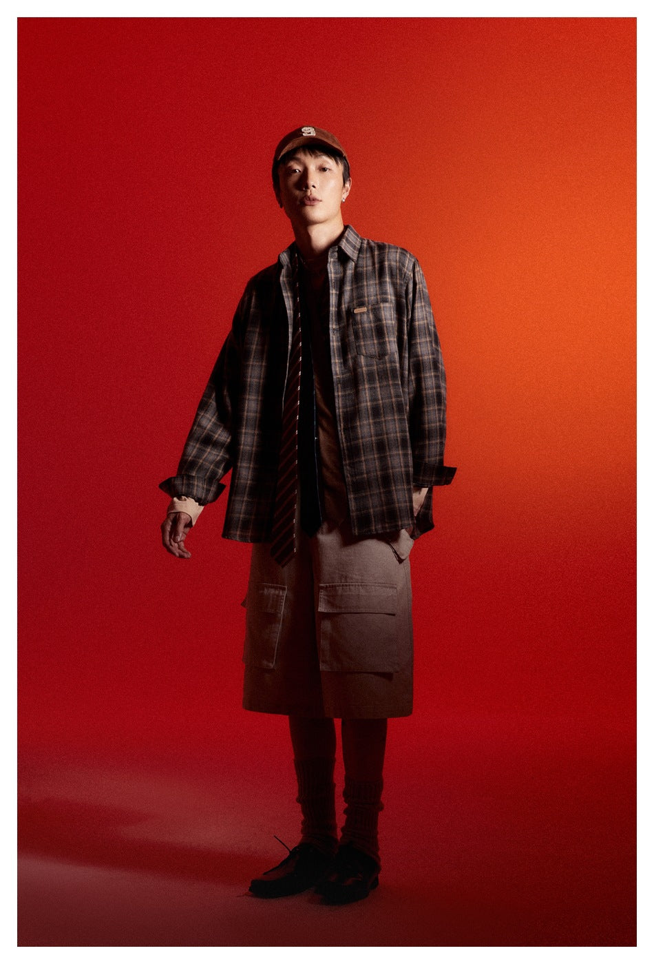 Man in an open gray plaid long-sleeve shirt layered over a tee with tie and cargo shorts, red studio background