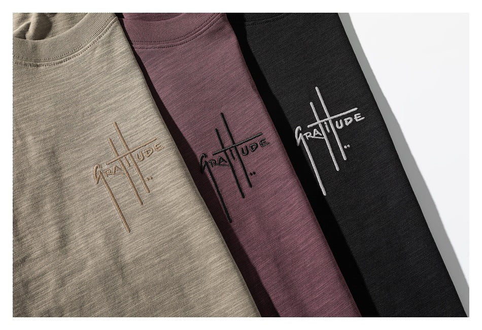 Three folded T-shirts in beige-gray, muted mauve, and black, each with embroidered lettering and line artwork.