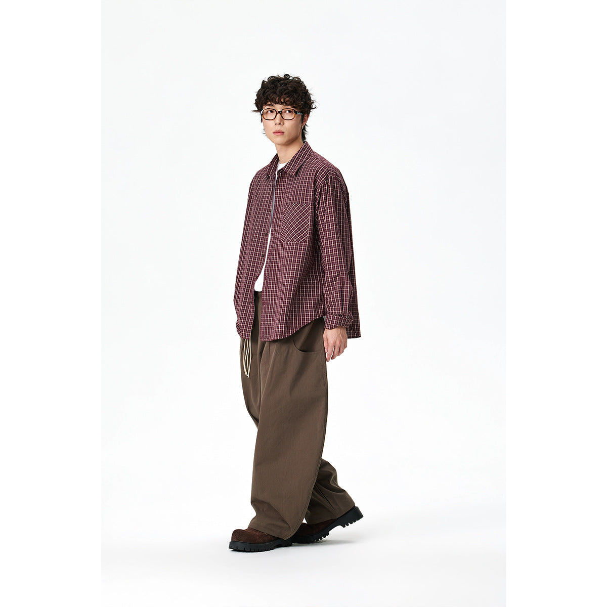 Full-length model wearing a burgundy plaid button-up shirt with a contrasting chest pocket and loose brown trousers.