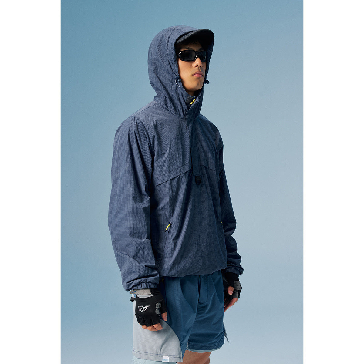 Model wearing a dark navy hooded pullover jacket with yellow zipper pulls, sunglasses, gloves and blue shorts.