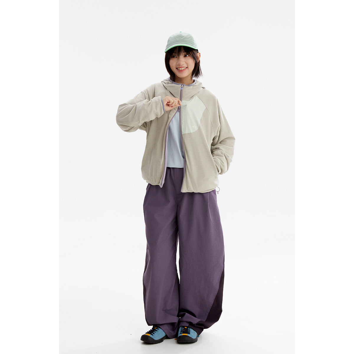 Model wearing a light beige hooded zip jacket with a pale chest panel and loose purple trousers.
