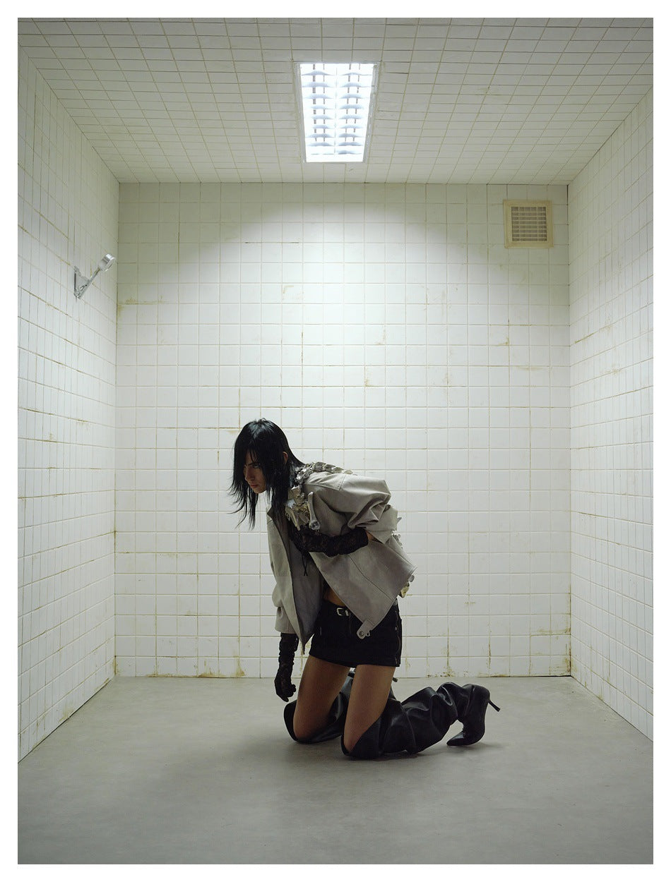 Model kneeling in a white tiled room wearing a gray jacket with black gloves, mini skirt and tall black boots