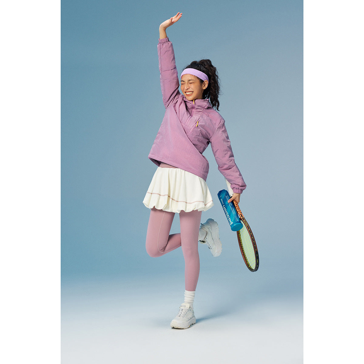 Woman in a lilac pullover jacket, white skirt and pink leggings poses with a tennis racket.