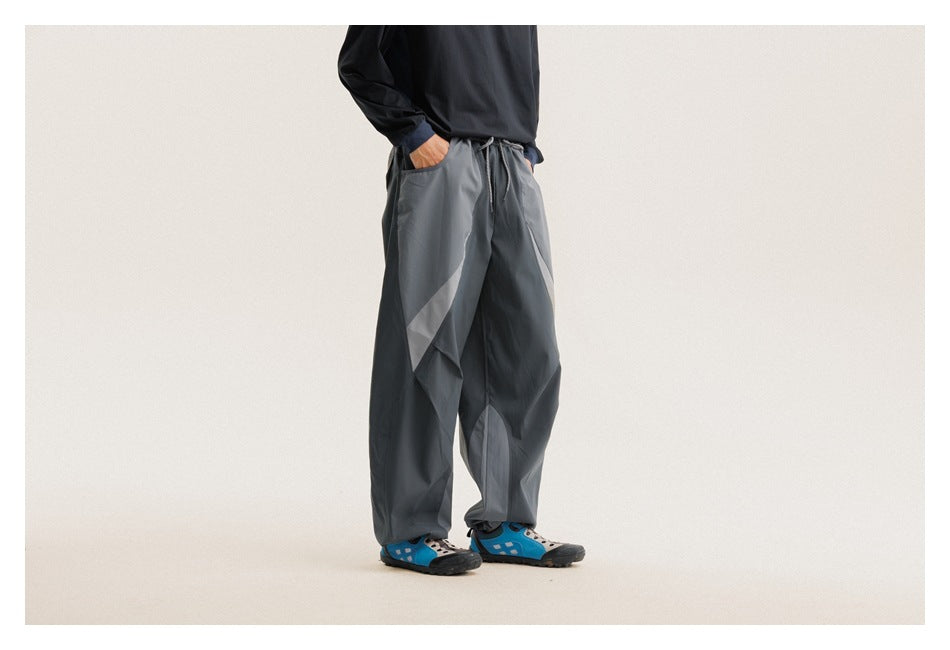 Front view of a model wearing loose charcoal-gray pants with a drawstring waist, side pockets and angular contrast panels.