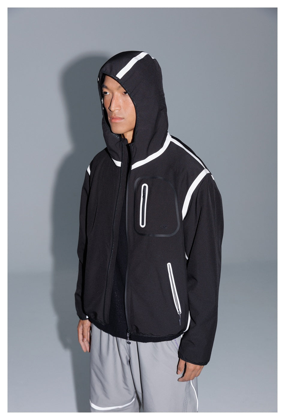 Model wearing a black hooded jacket with white piping and zip pockets, shown in a close front view.