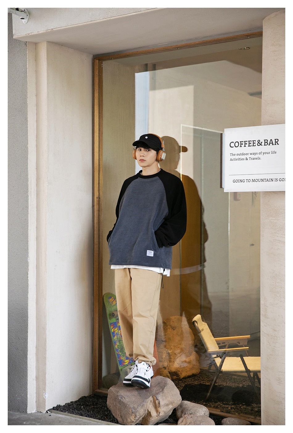 Model in a gray and black raglan sweatshirt with hands in pockets, tan pants and sneakers standing in a glass doorway