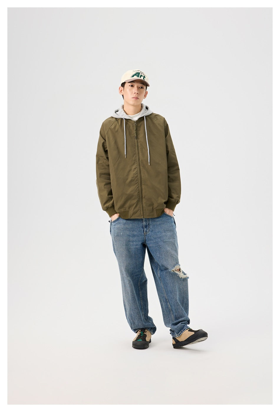 Full-length model wearing an olive jacket with a gray hood, loose blue jeans, and a beige cap.