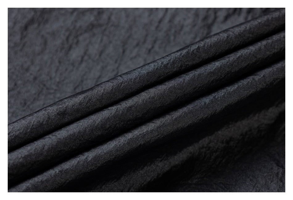 Close-up of dark charcoal-black fabric folded into diagonal ridges, showing its subtle sheen and texture.