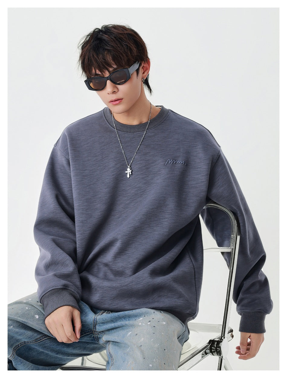 Model seated in a charcoal gray oversized crewneck sweatshirt with a small embroidered chest mark and ribbed cuffs.