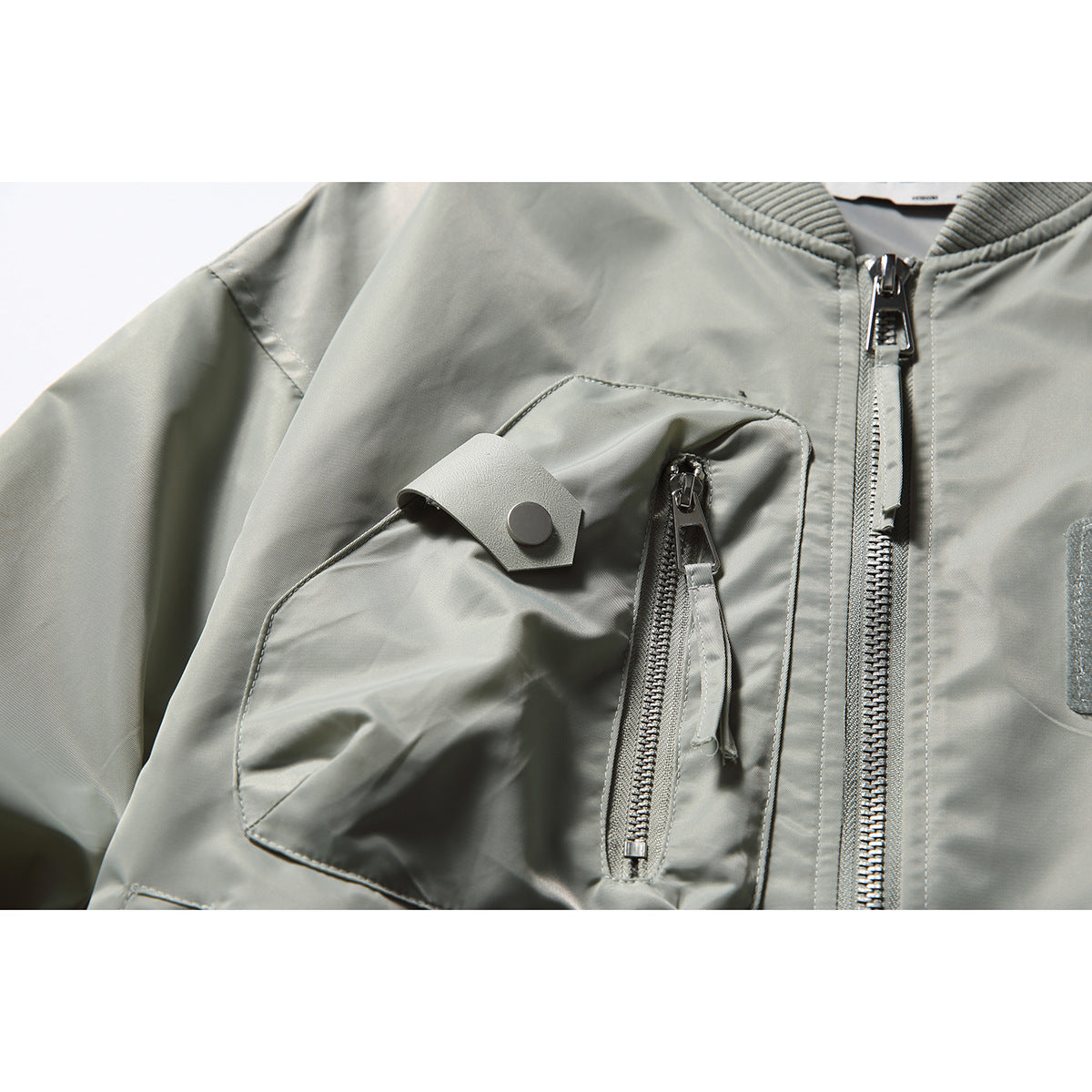 Close-up of a pale gray bomber jacket's zippered chest pocket, snap flap, and front zipper.