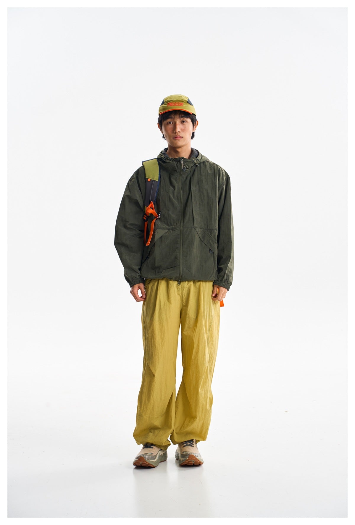 Front view of a model wearing a dark olive hooded jacket and loose mustard-yellow trousers against a white studio background.