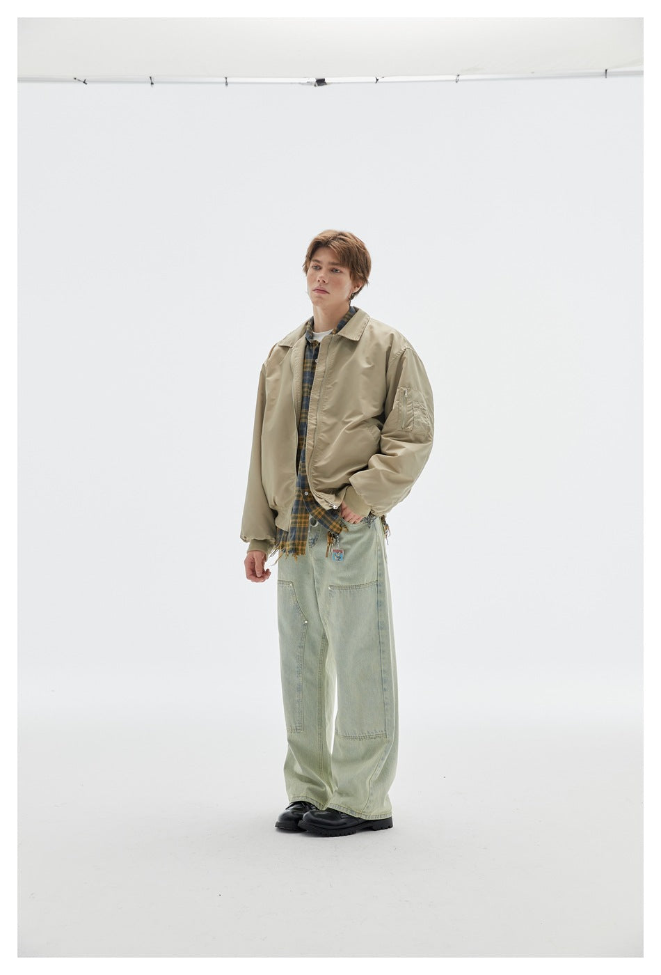 Full-length model wearing a beige bomber jacket with a plaid shirt, loose pale green jeans and black shoes.