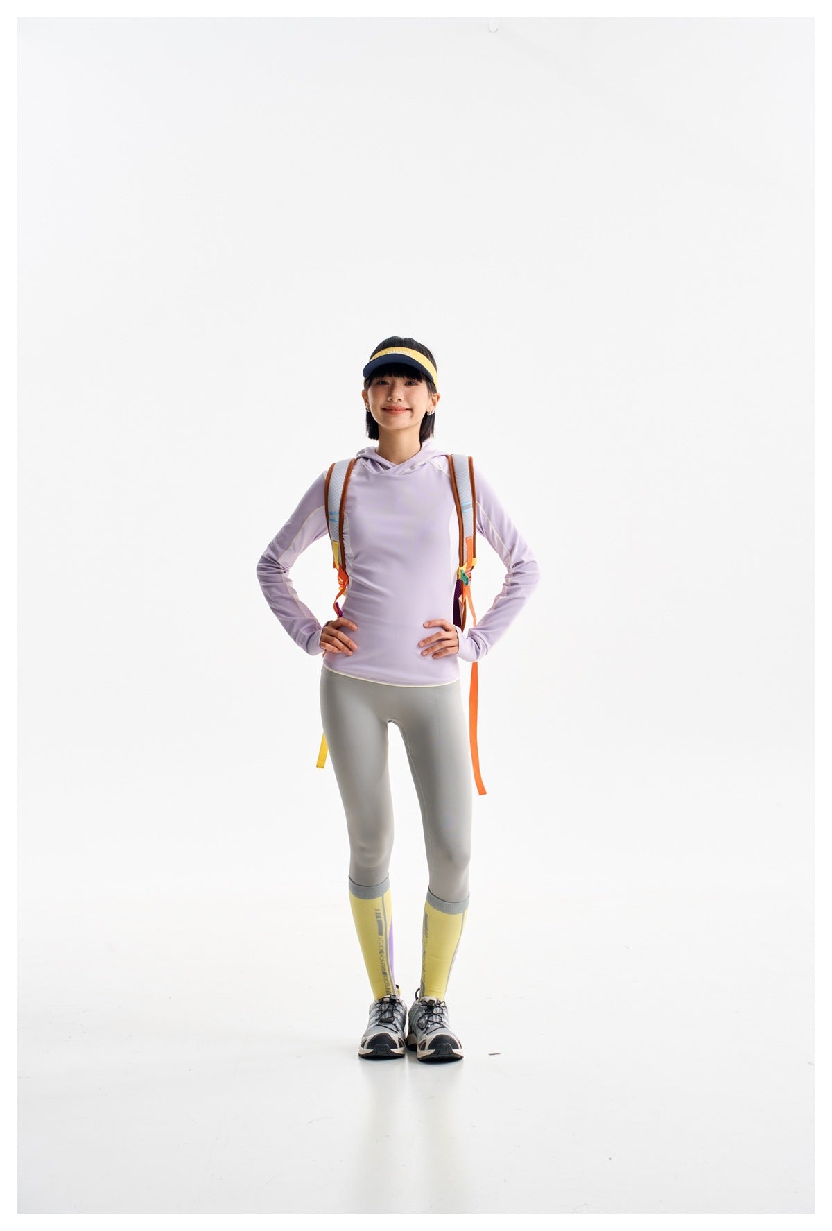 Full-length model wearing a lavender hoodie, gray leggings and yellow socks, with a colorful backpack and visor.