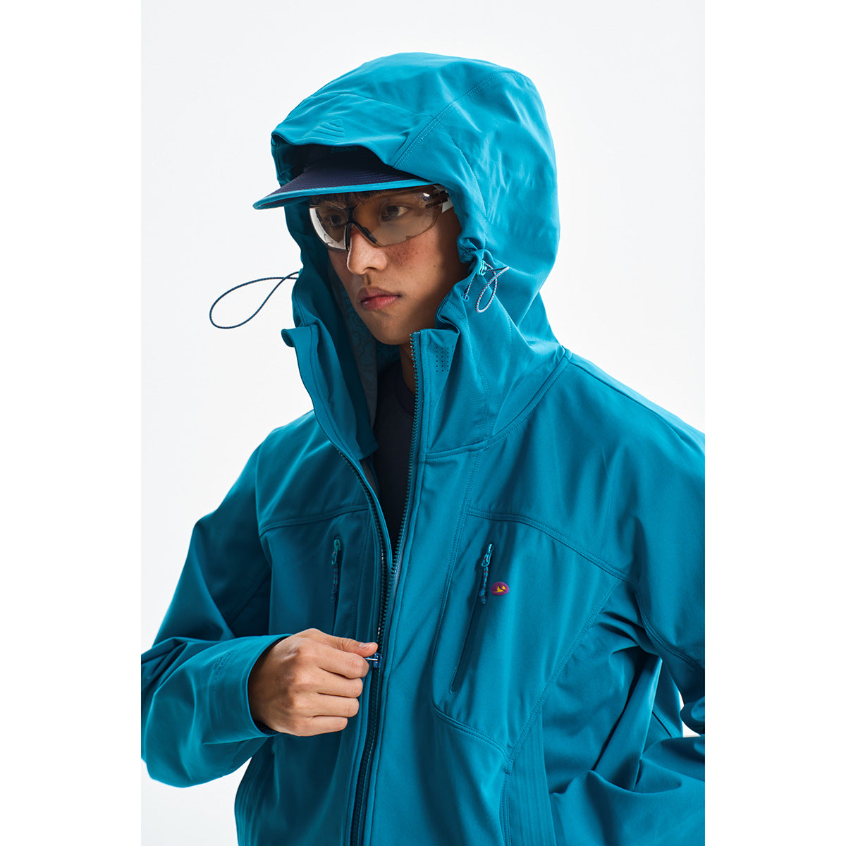 Close view of a model wearing a bright teal hooded jacket with a brimmed cap and tinted glasses.