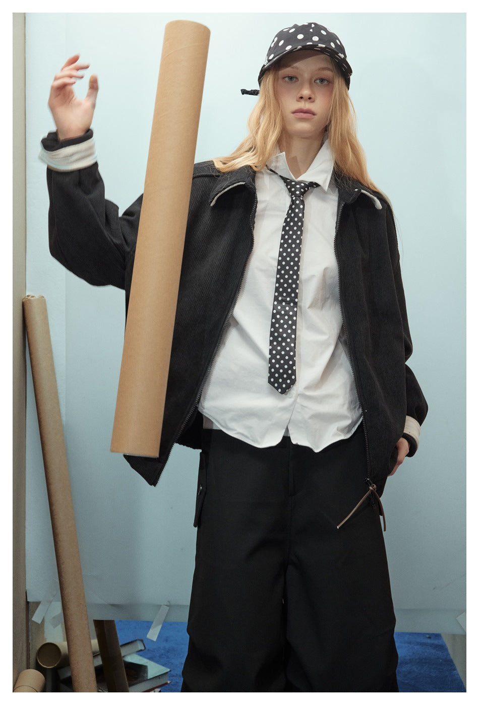 Model wearing an open black jacket with cream-trimmed cuffs over a white shirt and polka-dot tie and cap.