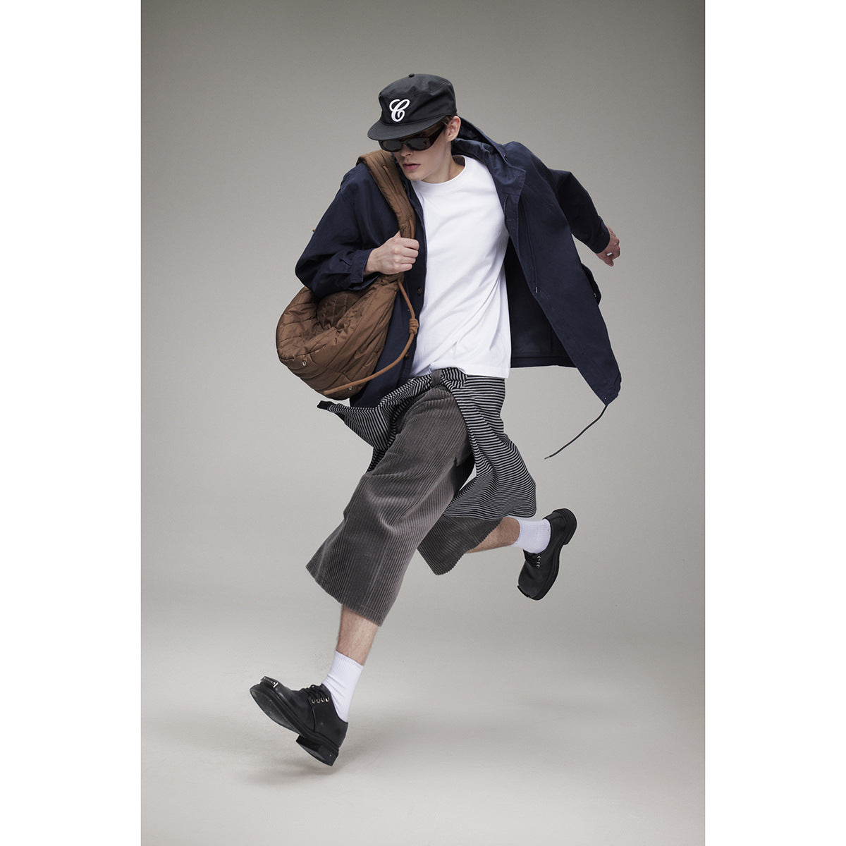 Model in a navy hooded jacket, white T-shirt, and loose gray cropped trousers, carrying a brown bag.