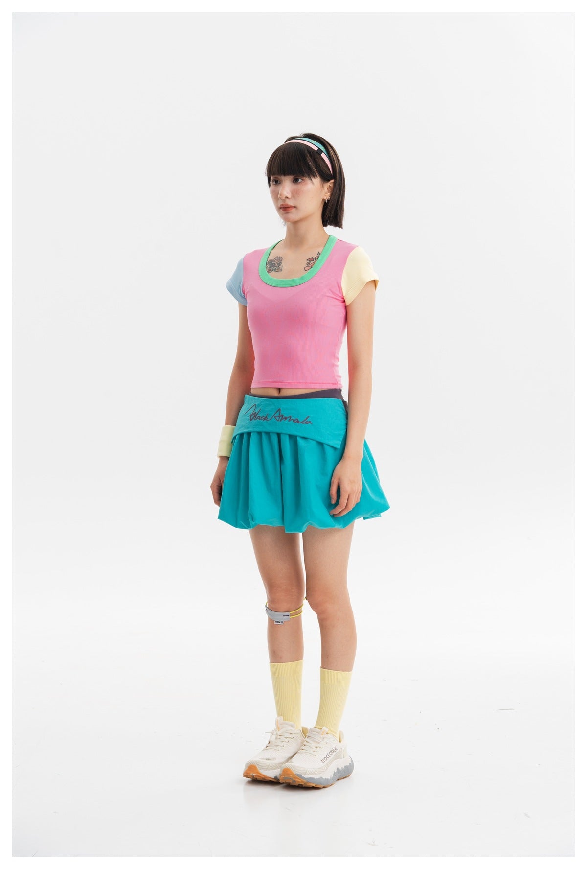 Model wearing a pink cropped ribbed tee with green neckline, blue and yellow sleeves, styled with a teal mini skirt