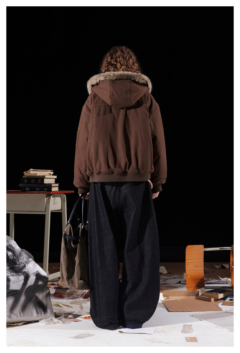Rear view of a model wearing a brown jacket with a fur-trimmed hood, gathered hem, and dark wide-leg trousers.