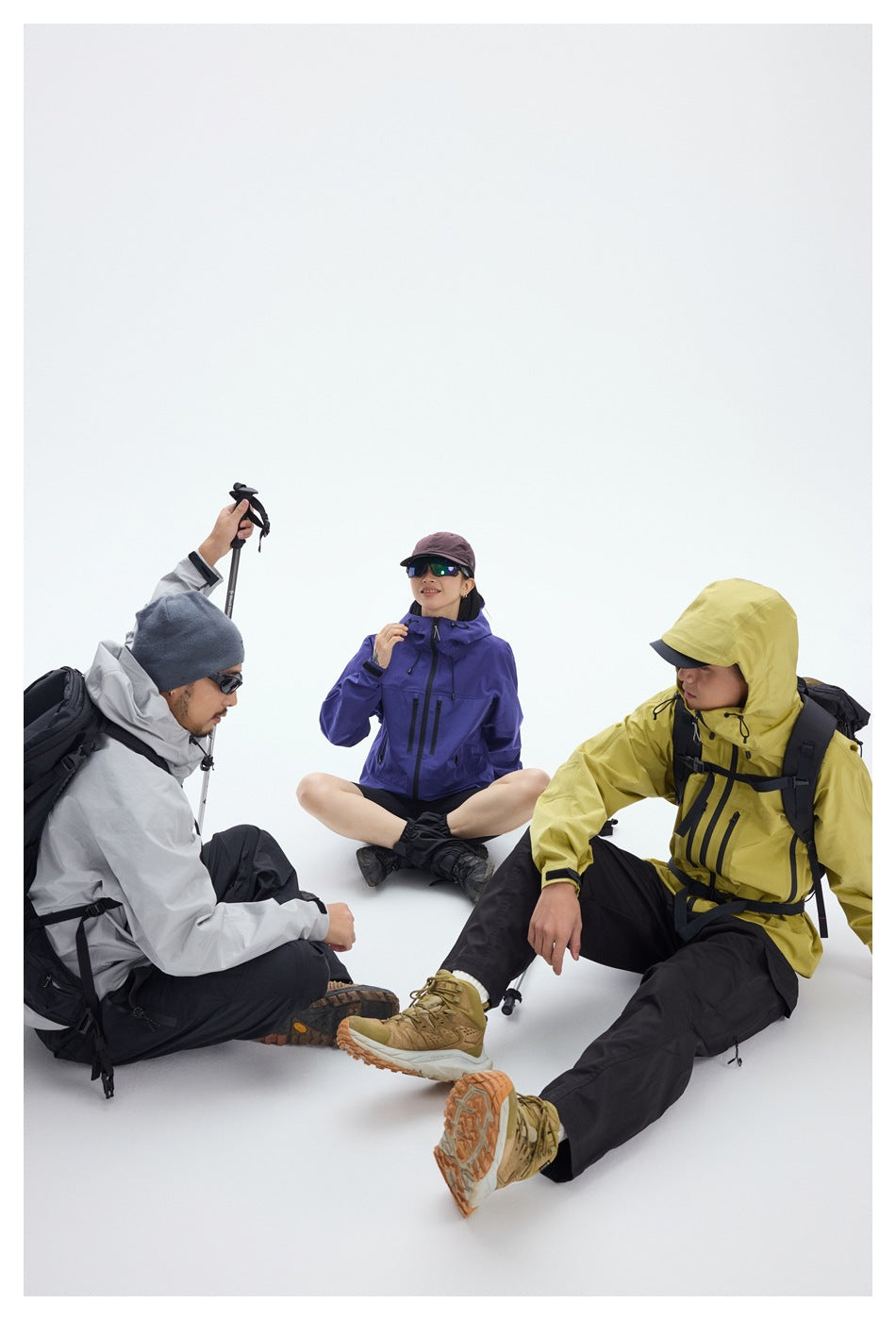Three hikers relax on the floor in gray, purple, and yellow hooded jackets with black pants.