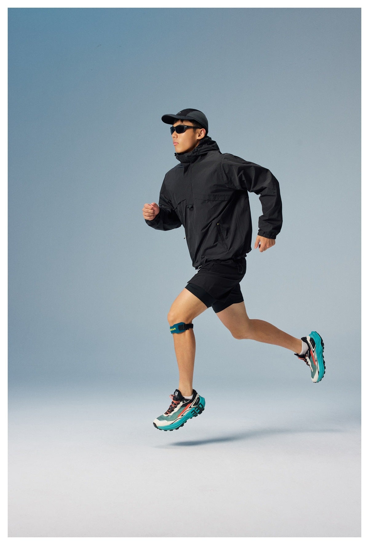 Man running in a black hooded windbreaker and shorts against a blue studio backdrop.
