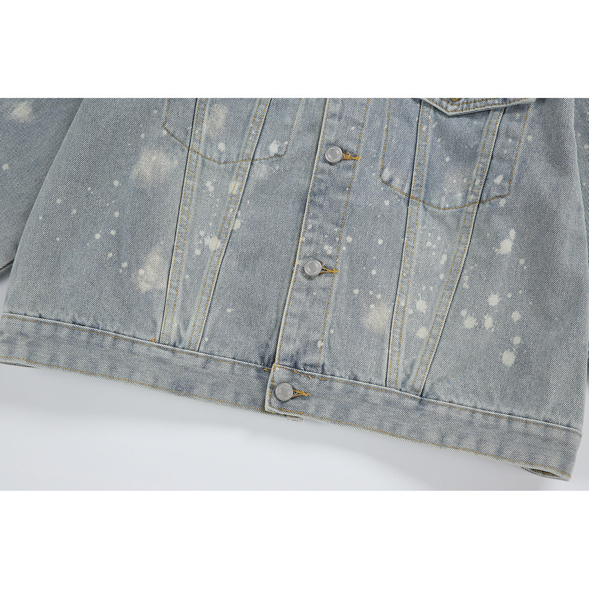Close-up of a light-wash denim jacket front with pale splatter marks, flap pockets, and metal buttons.