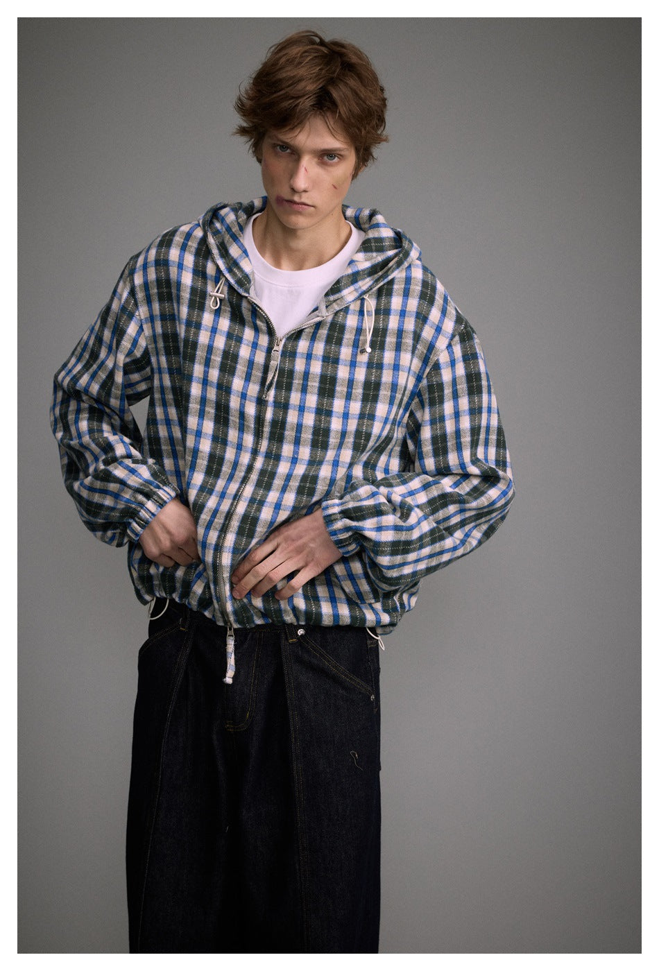 Model wearing a blue, cream, and black plaid zip-front hooded jacket with dark jeans.