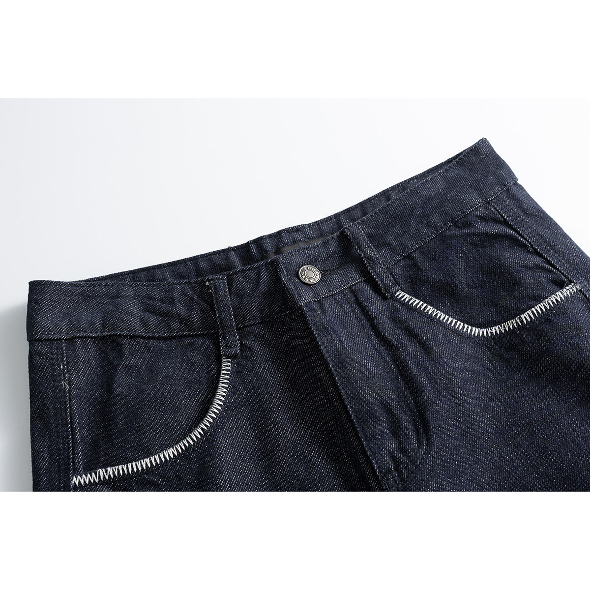 Close-up of dark indigo jeans showing the waistband, metal button, belt loop and curved front pockets with white stitching.