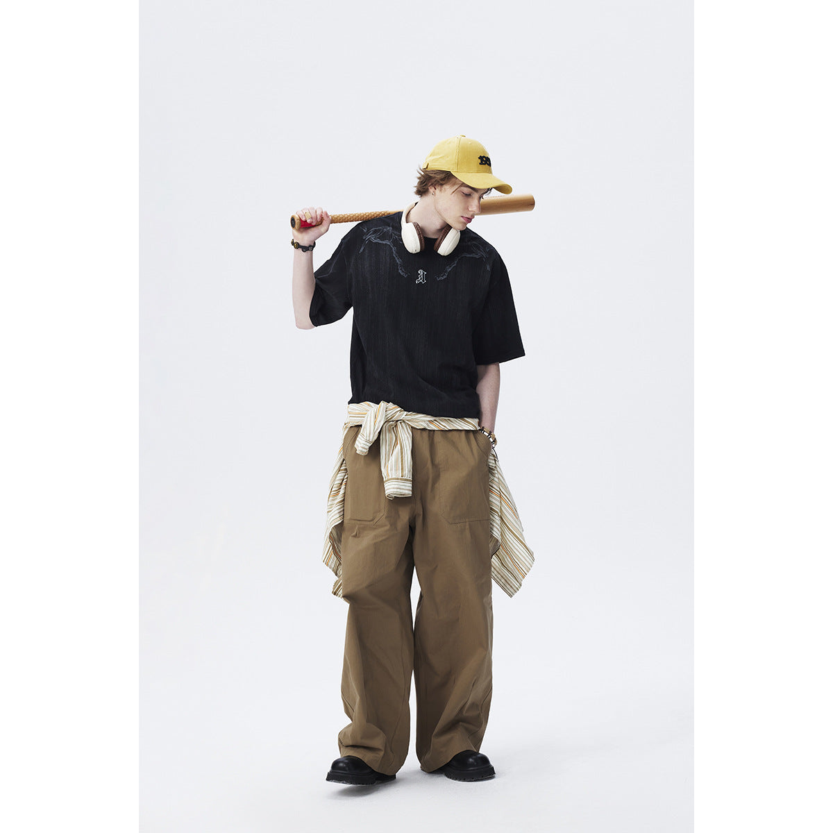 Model wearing a black graphic T-shirt and loose brown pants, with a yellow cap and striped shirt tied at the waist.