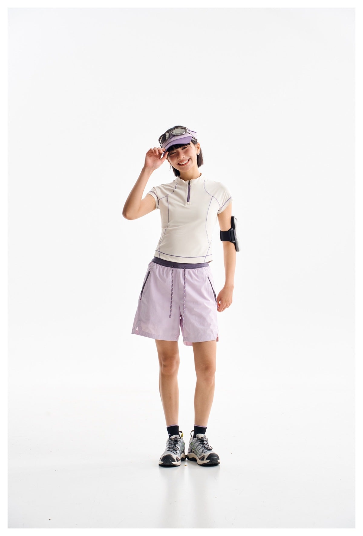 Full-length model wearing a white quarter-zip top with purple piping and lavender shorts, visor, and running shoes.