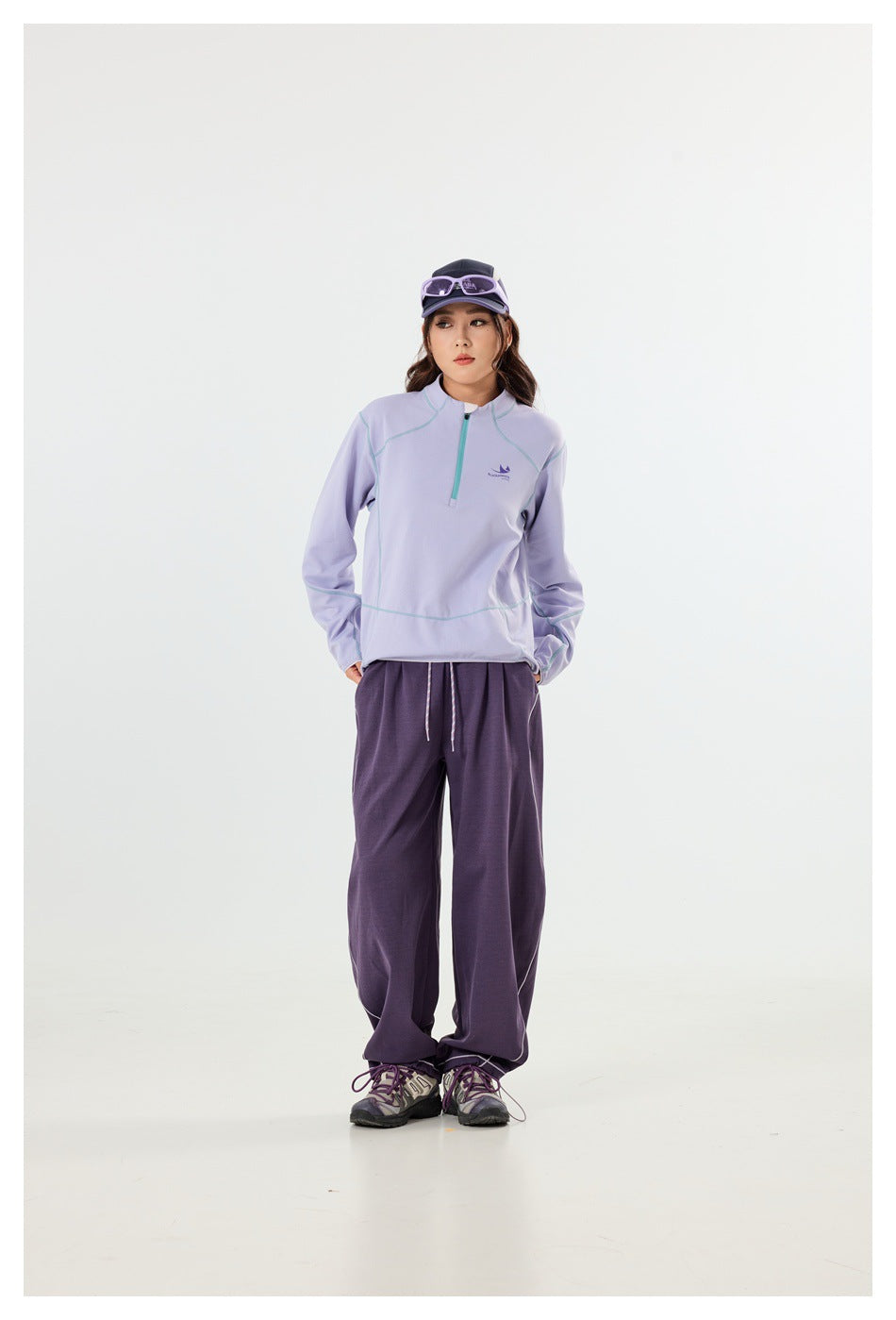 Front view of a model wearing a lavender quarter-zip pullover with pale contrast seams and loose purple pants.