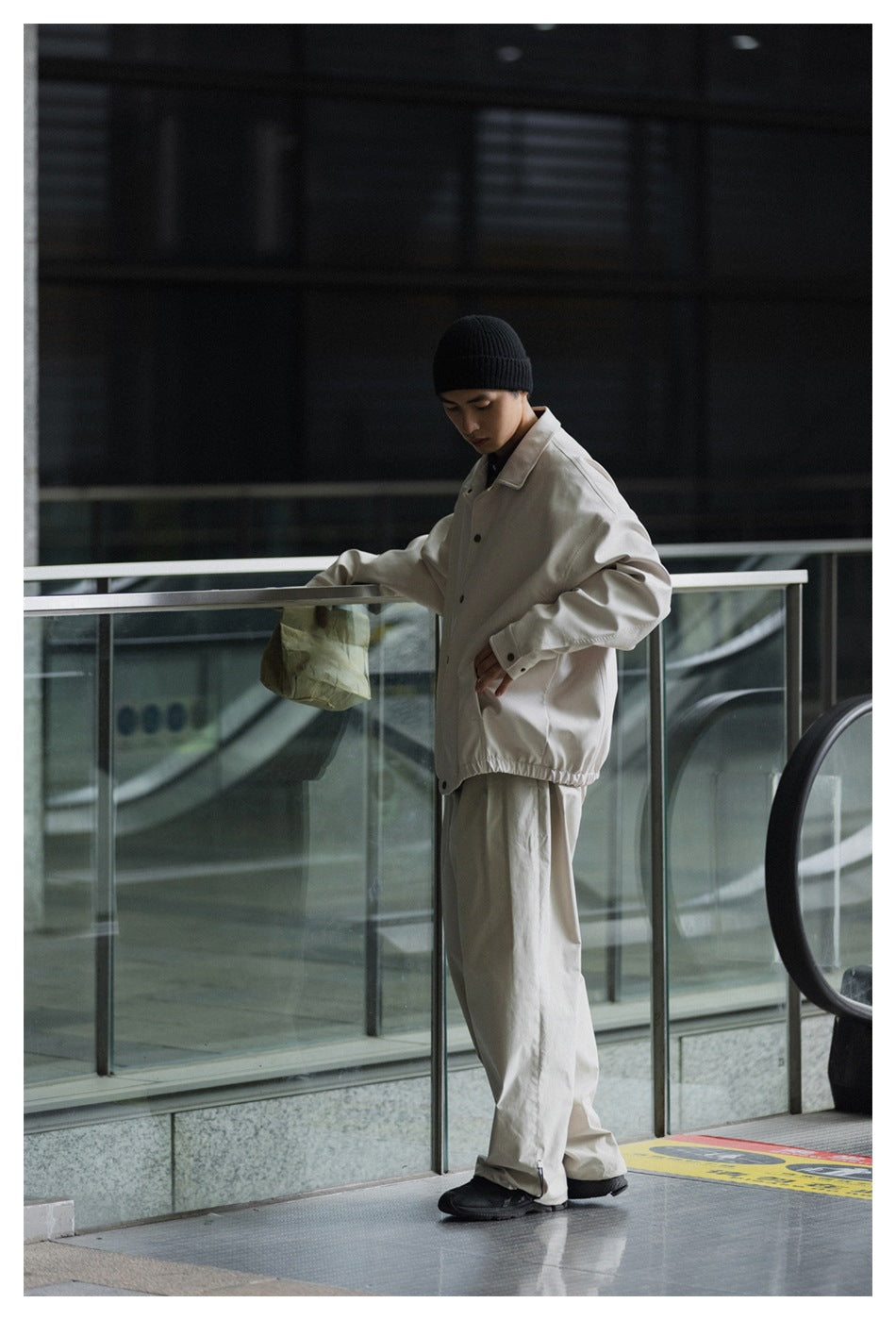 Model wearing a light beige button-front jacket with an elastic hem, loose matching trousers, and a black beanie.