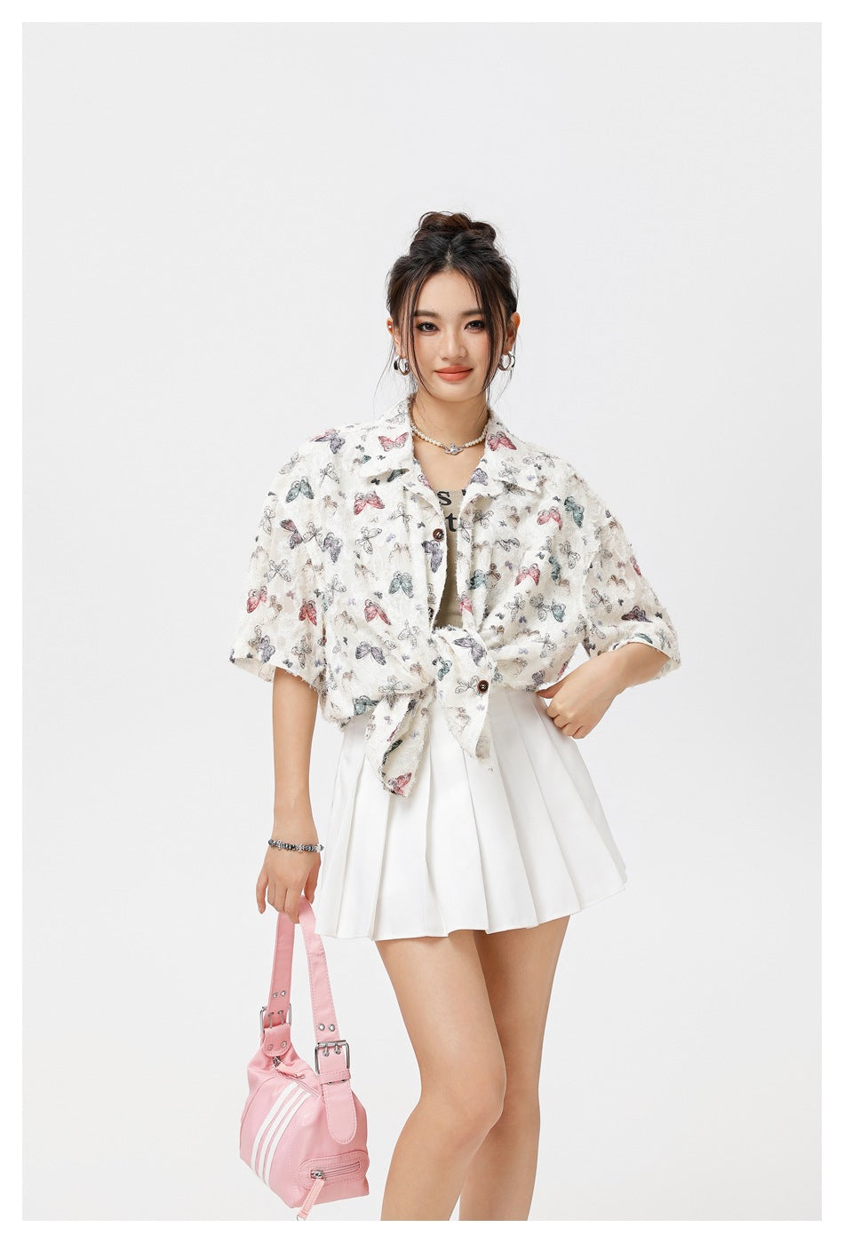 Model wearing a cream butterfly-print short-sleeve shirt tied at the waist over a white pleated skirt.