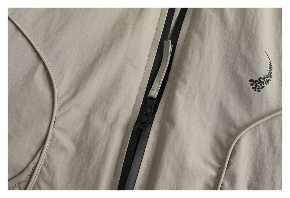 Close-up of a light taupe jacket with a black front zipper, curved seam, and small dark abstract chest motif.