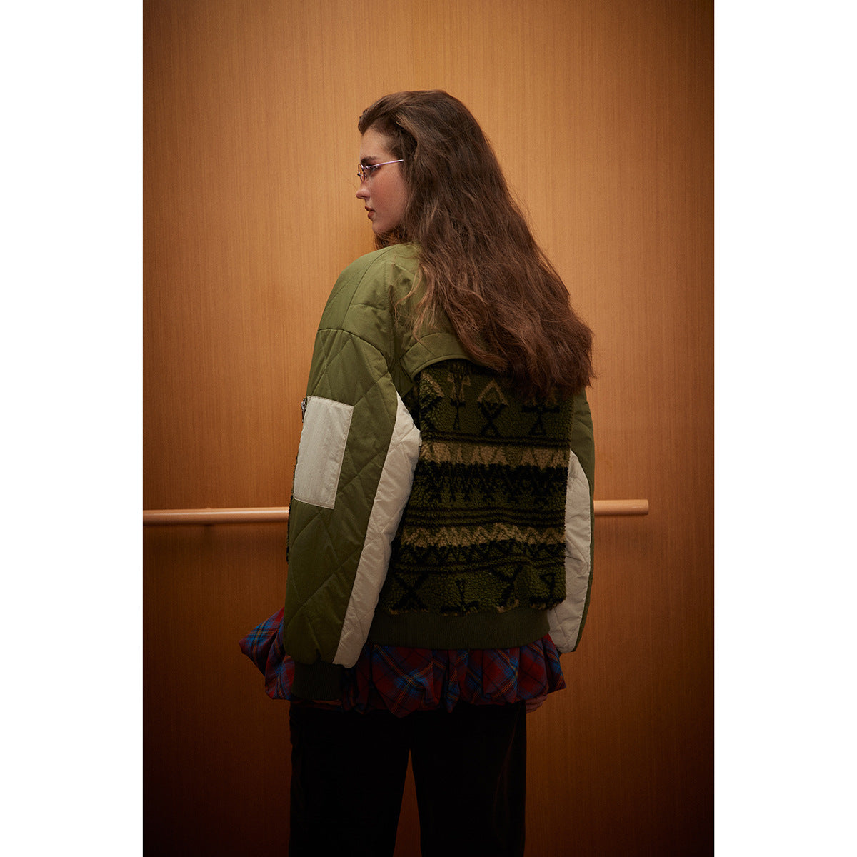 Back view of a model wearing an olive bomber jacket with quilted shoulders, cream sleeve panels, and a dark patterned back.