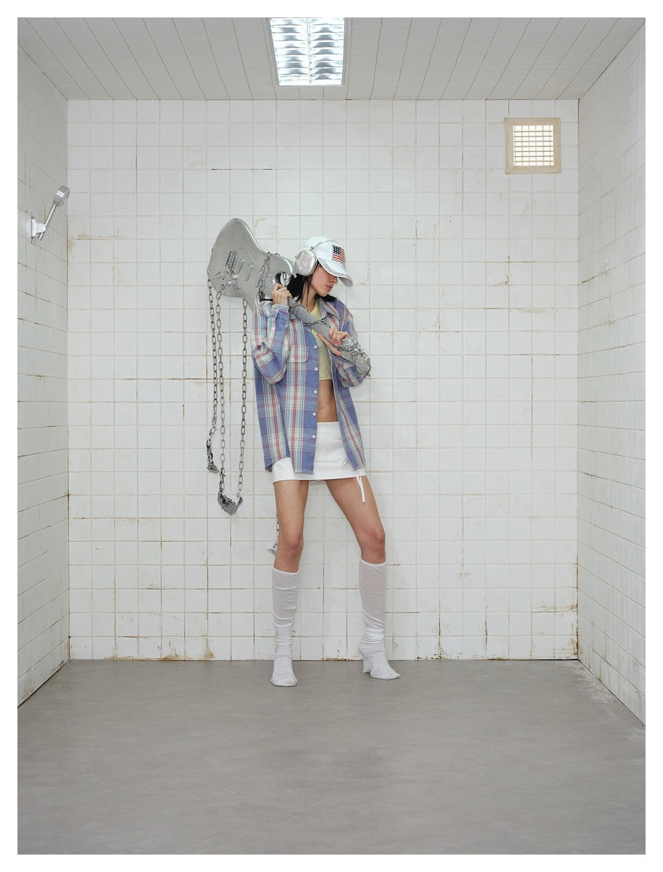Model in an oversized blue plaid shirt over a pale yellow top and white skirt, with a white cap and silver earmuffs.