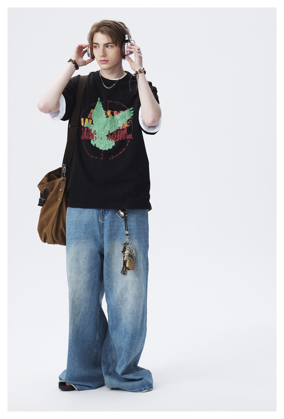 Model wearing a black graphic T-shirt over white sleeves, with a raised green bird design and loose blue jeans.