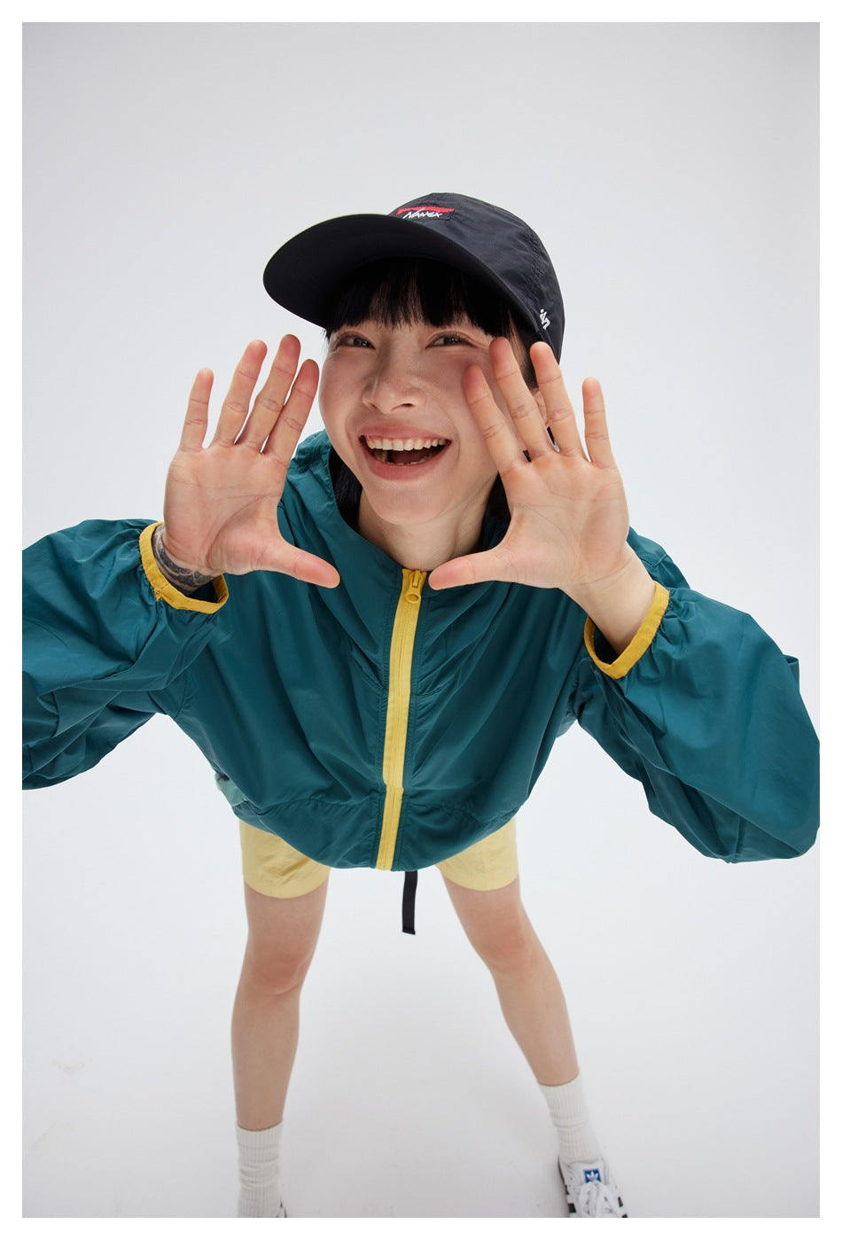 Model wearing a teal hooded jacket with a yellow zipper and cuffs, leaning toward the camera.