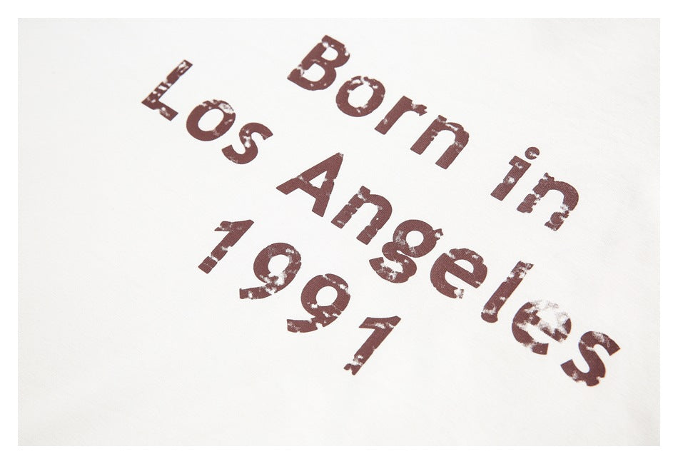 Close-up of distressed brown “Born in Los Angeles 1991” lettering printed on a white T-shirt.