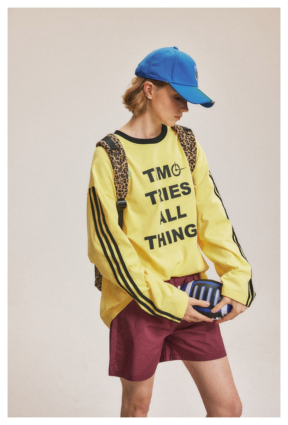 Three-quarter view of a model in a yellow striped slogan sweatshirt and burgundy shorts, holding a ball.