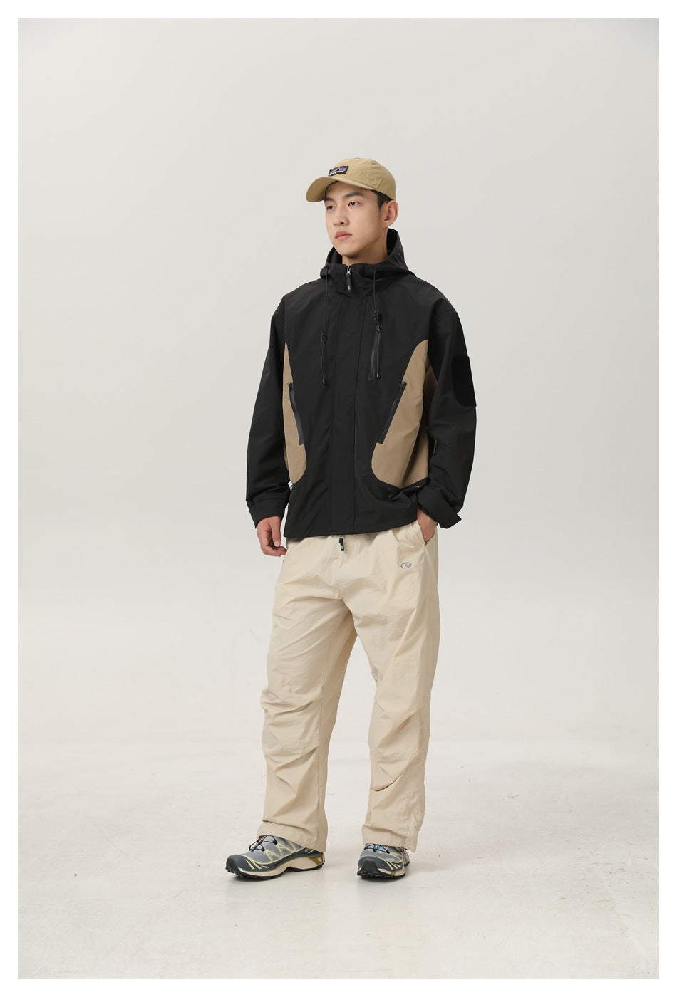 Full-length model wearing a black and tan hooded jacket with beige trousers and a tan cap.