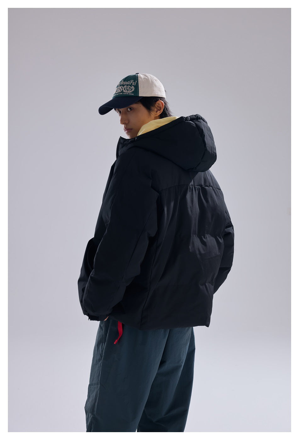 Model in a black hooded puffer jacket, turned to show the back and side against a gray studio background.