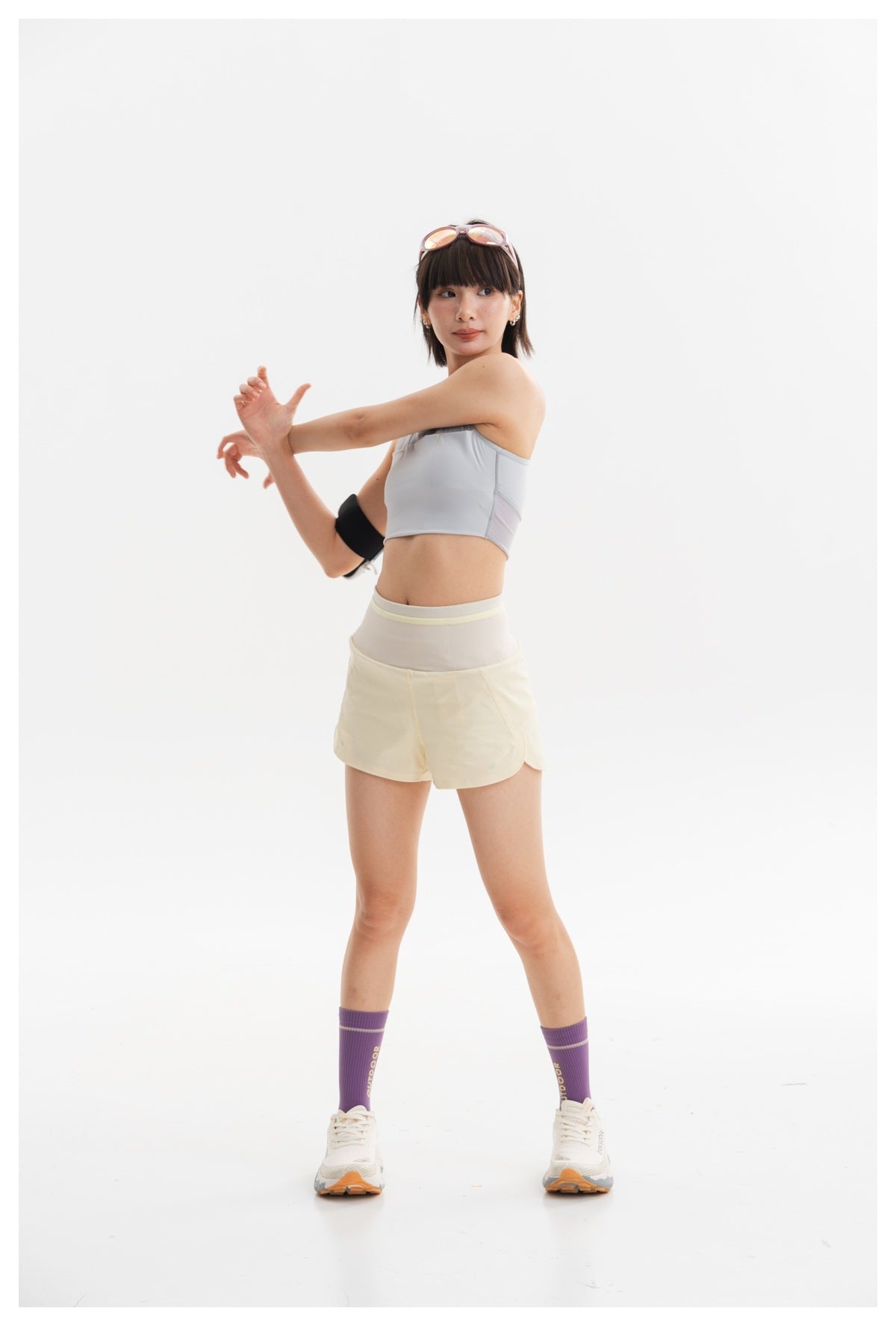Model stretching in a pale gray cropped athletic top and cream running shorts, shown full length against a white background.