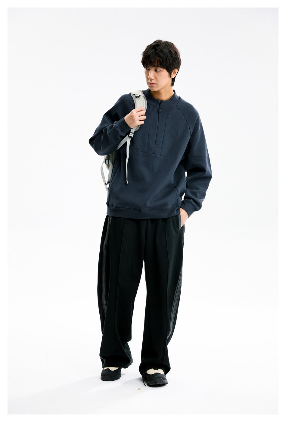 Full-length three-quarter view of a model wearing a dark navy pullover, black wide-leg pants, and a light backpack.