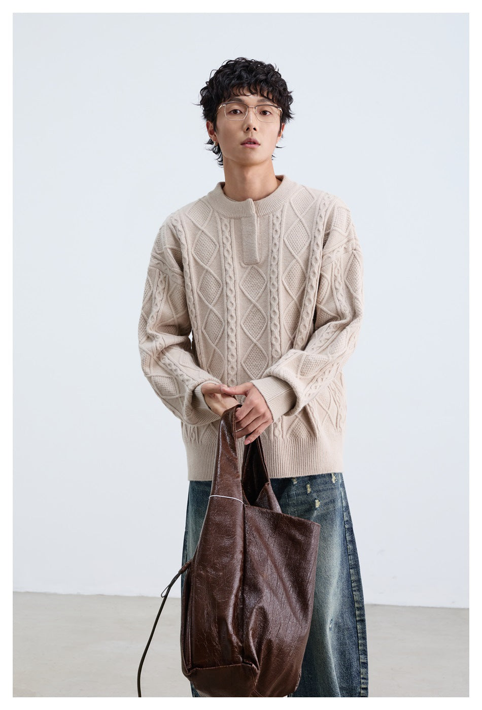 Front view of a model wearing a cream cable-knit sweater with raised diamond and braided patterns, holding a dark tote.