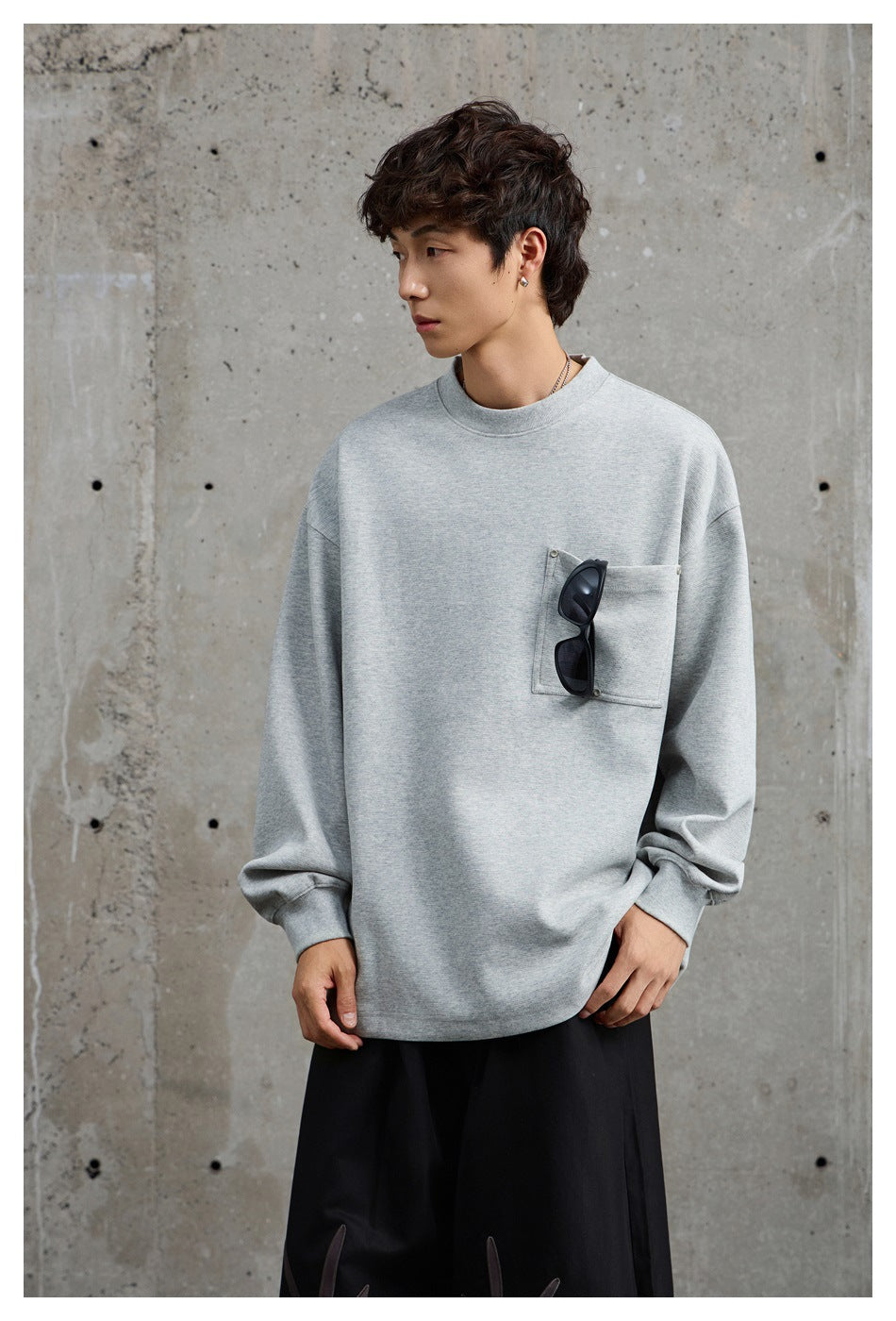 Model wearing a gray crew neck sweatshirt with chest pocket, sunglasses hooked on the pocket, against a concrete wall