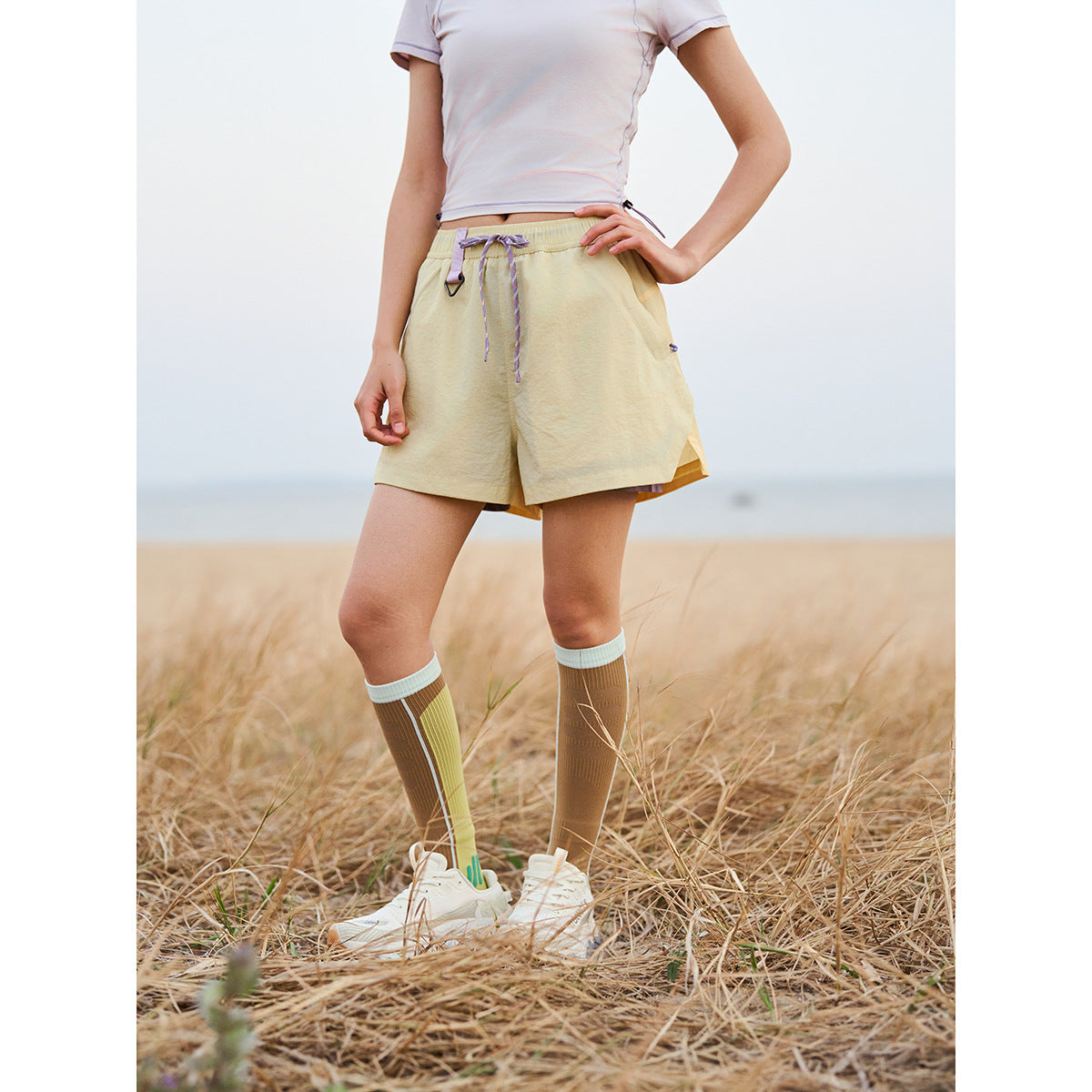 Cropped model wearing pale yellow drawstring shorts with side pockets, striped knee socks and white sneakers outdoors.