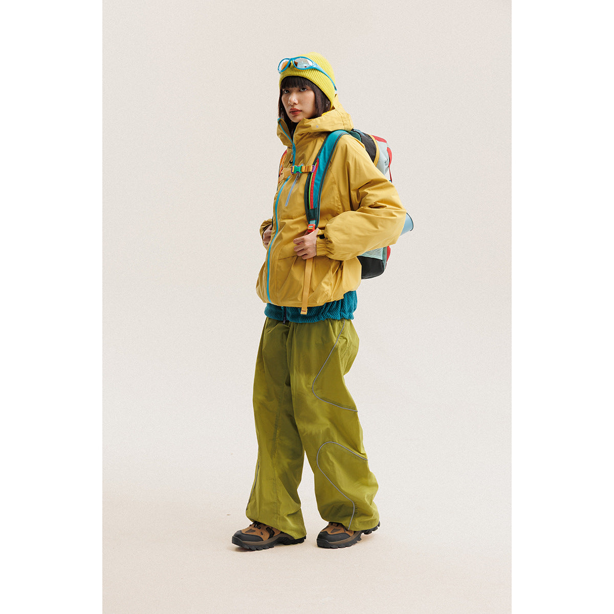 Model wearing a mustard hooded jacket and olive trousers with a colorful hiking backpack.